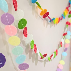 May include: A colourful felt garland with circles in various colours, including pink, yellow, green, red, and purple. The garland is strung together with white thread.
