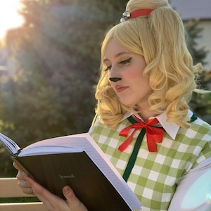 May include: A person in a cosplay costume, reading a book. The costume features a blonde wig with a red headband and a bell, a green and white checked vest, and a red bow tie. The book is open, with the word "Journal" visible.