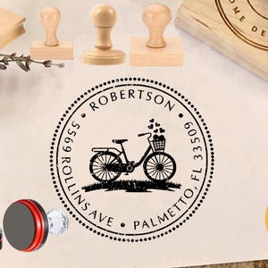 Może przedstawiać: Kolekcja drewnianych stempli i przezroczysty stempel z czerwoną i czarną podstawą. Główny stempel przedstawia rower z koszem pełnym serc, otoczony tekstem, w tym imieniem, adresem i słowem "Palmetto, FL".