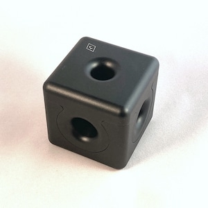 Puede incluir: Un cubo de rompecabezas negro con un agujero en el centro de cada lado. El cubo tiene un pequeño símbolo de flecha en la parte superior.