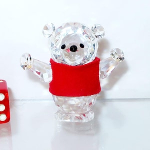 May include: A crystal bear figurine with a red vest and outstretched arms. The bear has a black nose and eyes. A red die with white dots is next to the bear.