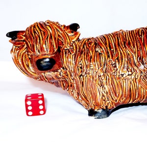 May include: A ceramic Highland cow figurine with a textured, brown and tan coat. The figurine has black horns and hooves. A red die with white dots is next to the cow.