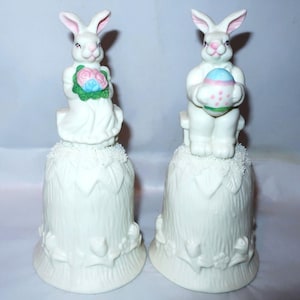 May include: Two white ceramic Easter bunny figurines. One bunny holds a bouquet of pink and blue flowers, the other holds a decorated Easter egg. Both bunnies sit atop white bell-shaped bases with floral and leaf details.