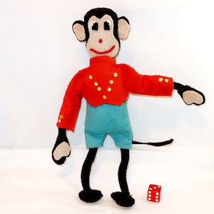 May include: A felt monkey doll with a black head, ears, arms, and legs, featuring a beige face, red nose, and red mouth. The doll wears a bright red jacket with yellow dots and light blue shorts. A small red six-sided die sits on the floor.