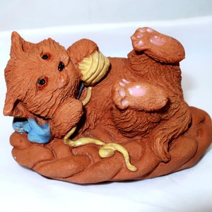 May include: A brown ceramic sculpture of a playful cat lying on its back, paws up, with a ball of yarn. The cat has three eyes and pink paw pads. A blue object is near the cat's mouth. The sculpture is on a brown base.