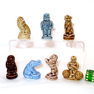 May include: A collection of ceramic figurines, including a lion, a man in a suit, a tiger, a monkey, an elephant, a bear, and a strongman holding weights. A green die is also present. The figurines are in various colors, including brown, blue, and tan.