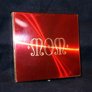 May include: A red, square compact with the word "MOM" in an elegant, gold script. The compact has a brushed metal finish and a gold trim. The background is a dark, solid color.