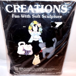 May include: A Creations Fun With Soft Sculpture craft kit package featuring a white bunny wreath, a lavender cat, a blue cat, and a small cream-colored cat. The black plastic packaging displays the product name, item numbers, and contents list.