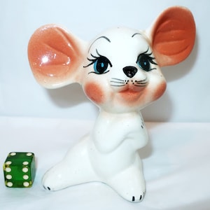 May include: A vintage ceramic mouse figurine with large ears, rosy cheeks, and blue eyes. The white mouse is posed in a seated position. A green die with white dots is next to the mouse.