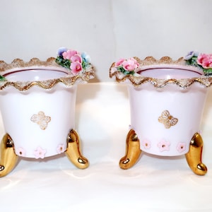 May include: Two matching pink ceramic planters with gold accents. Each planter features a scalloped rim with floral embellishments, a gold butterfly design, and gold-colored feet. The planters are approximately 4 inches tall.