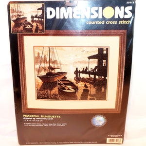 May include: A Dimensions counted cross stitch kit titled Peaceful Silhouette, designed by James Himsworth. The packaging features a sepia-toned scene of boats docked at a pier at sunset. The finished design measures 13 inches by 10 inches.