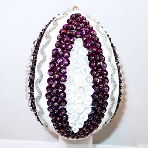 May include: A decorative egg covered in white, purple, and silver sequins. The center features a vertical oval pattern of white sequins bordered by dark purple sequins. Wavy silver trim lines the sides, with additional purple and clear beads.