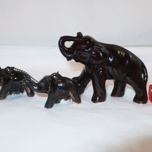 May include: Three dark brown ceramic elephant figurines, one large and two smaller, are arranged on a white surface. A red die with white dots is next to the elephants. The elephants have a glossy finish.