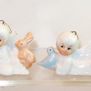 May include: Four white ceramic angel ornaments with gold hanging loops. Each angel features a different animal: a squirrel, a rabbit, a blue bird, and a goose. A small red die with white dots sits to the right of the figures for scale reference.