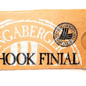 May include: A tan Longaberger Foundry Collection box with the words "HOOK FINIAL" in black. A green die with white dots is next to the box. The box has a circular logo.