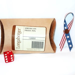 May include: A brown Longaberger box with a label that reads "AMERICAN PRIDE PIN" and the number "12655". A red die with white dots and a ribbon pin in the colors of the American flag are also present.