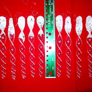 May include: A collection of clear glass icicle ornaments with a spiral design. Each ornament has a frosted top with glitter and sequins. The ornaments are arranged on a red surface, with a ruler for scale.