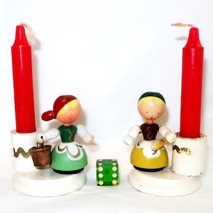 May include: Two red candles in white holders flank two wooden figurines, one holding a bucket. A green die with white dots sits between them. The figurines wear green and yellow dresses.