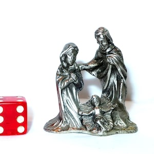 May include: A silver-colored nativity scene figurine depicting Mary, Joseph, and baby Jesus. A red die with white dots is next to the figurine. The figurine is on a white background.
