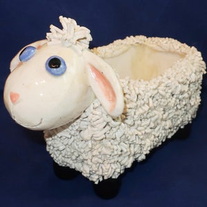 May include: A white ceramic sheep planter with a textured woolly body, pink-lined ears, and blue eyes. The sheep has a smiling face and sits on four black feet. The planter's opening is at the top, ready for plants.