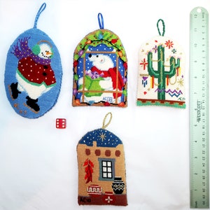May include: Four handmade needlepoint Christmas ornaments. Designs include a snowman, a polar bear, a cactus, and a Southwestern-style building. Each ornament has a loop for hanging. A red die and a ruler are also in the image.