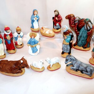 May include: A collection of hand-painted ceramic nativity scene figurines. The set includes figures of Mary, Joseph, the Three Wise Men, an angel, baby Jesus, animals, and other characters. The figurines are painted in various colors.