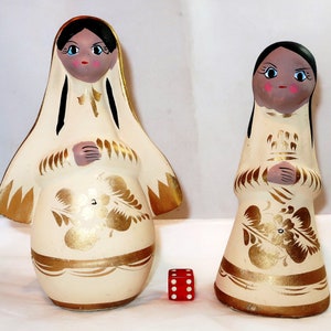May include: Two ceramic figurines of women in cream-colored dresses with gold accents and floral designs. The figurines have black hair, blue eyes, and pink cheeks. A red die is next to the figurines. The figurines are approximately 6 inches tall.