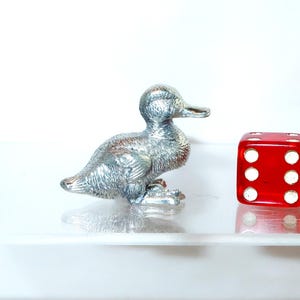 May include: A small, silver-colored duck figurine next to a bright red die with white dots. The duck has detailed feather textures and is positioned facing right. The die shows the number six.