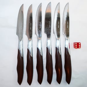 May include: Set of six steak knives with stainless steel blades and dark brown wooden handles. The knives have a silver bolster and are labeled "JAPAN SWORD". A red die with white dots is next to the knives.