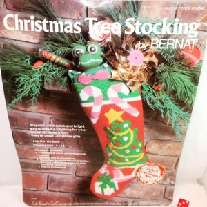May include: A Bernat Christmas Tree Stocking needlepoint kit package featuring a red stocking with a green tree, candy canes, and a frog character. The packaging includes text, product code WN8008, and states a finished size of 9 by 16 inches.
