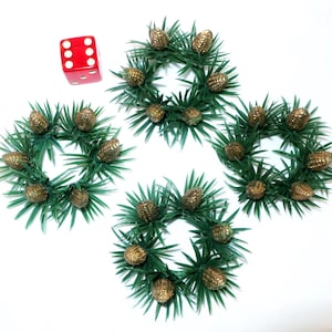 May include: Four small, green, artificial wreaths with gold-colored pinecones. A red die with white dots is next to the wreaths. The wreaths are arranged in a cross shape against a white background.