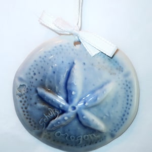 May include: A ceramic sand dollar ornament in shades of blue, with a white ribbon for hanging. The ornament features a detailed starfish design and the words "Schulz Oregon" inscribed on the surface. A decorative coastal-themed item.