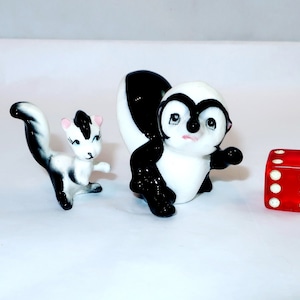 May include: Two ceramic skunk figurines, one small and one larger, in black and white. A red die with white dots is next to the larger skunk. The skunks have painted details, including pink ears and blue eyes.