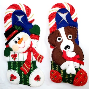 May include: Two Christmas stockings with felt appliqués. One features a snowman with a black hat, red scarf, and green mittens. The other has a brown and white dog with a red bow. Both stockings have green sequined sections and candy cane tops.