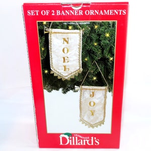 May include: A red box displays two banner ornaments. The banners are white with gold trim and lettering that spells "NOEL" and "JOY". The box reads "SET OF 2 BANNER ORNAMENTS" and features the Dillard's logo.
