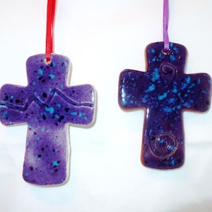 May include: Two ceramic crosses with a speckled purple glaze and blue accents. One cross has a mountain design, the other a spiral. Each is suspended by a ribbon, one red, one purple. Religious themed decor.
