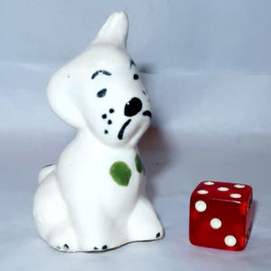 May include: A white ceramic dog figurine with black spots and a green spot on its side. Beside the dog is a red translucent die with white dots. The dog is sitting with its head tilted.