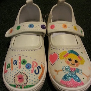 May include: A pair of white canvas shoes with a purple stitched design. The shoes are decorated with colorful buttons, a pink heart, and a hand-painted Lalaloopsy doll with glitter. One shoe says "Lalaloopsy" in colorful letters.