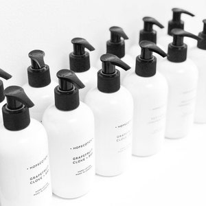May include: A row of white bottles with black pump tops and labels that read "Hopscotch Grapefruit Clove + Bay" hand lotion made in London.