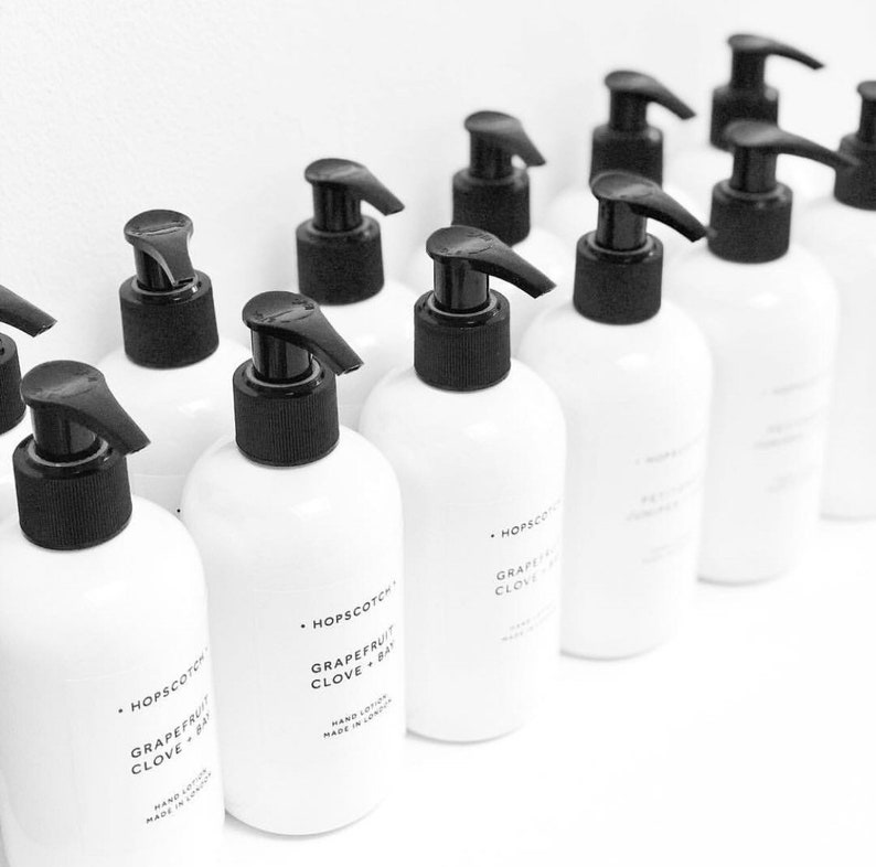 May include: A row of white bottles with black pump tops and labels that read "Hopscotch Grapefruit Clove + Bay" hand lotion made in London.