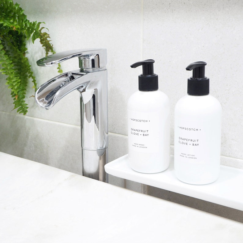 May include: Two white bottles of hand wash and hand lotion with black pump tops. The bottles are labeled "Hopscotch" and "Grapefruit Clove + Bay". The bottles are on a white shelf in a bathroom.