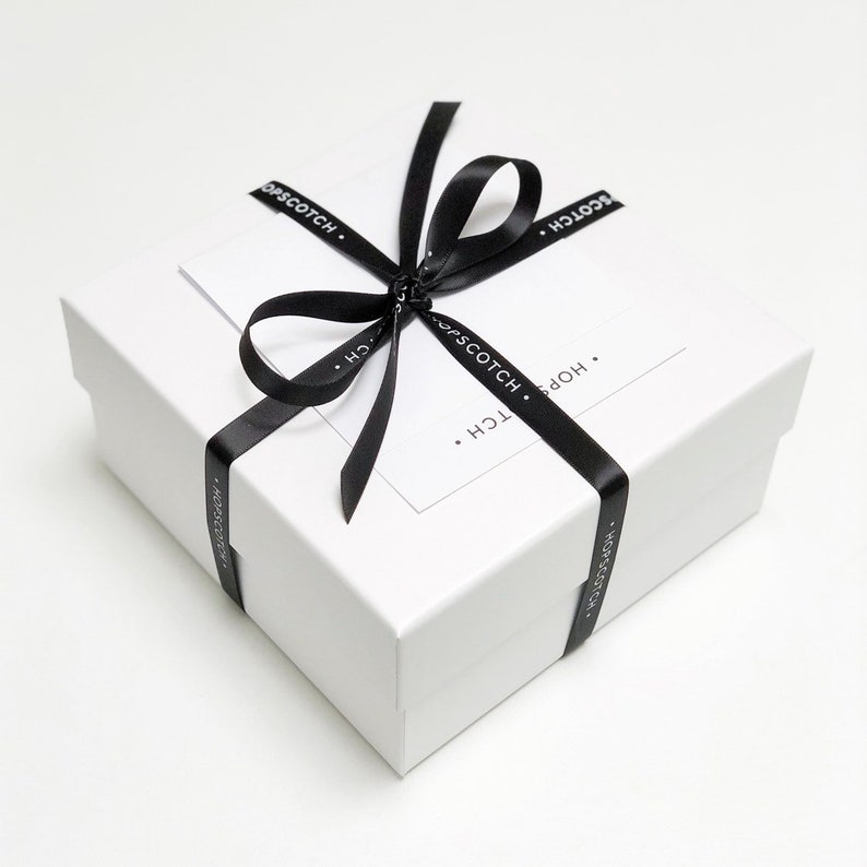 May include: A white gift box with a black ribbon tied in a bow. The ribbon has the text "HOPSCOTCH." printed on it.