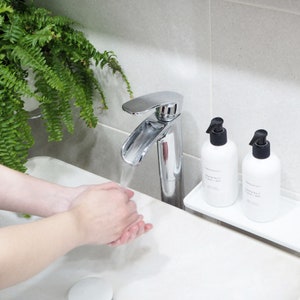 May include: A white bathroom sink with a chrome faucet and running water. Two white bottles of hand soap with black pumps are on a white shelf above the sink. The bottles have the text "Grapefruit & Bay" on them.