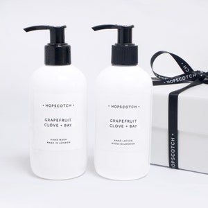 May include: Two white bottles of hand soap and lotion with black pumps. The bottles are labeled "HOPSCOTCH" and "GRAPEFRUIT CLOVE + BAY". One bottle is labeled "HAND WASH" and the other is labeled "HAND LOTION". Both bottles are made in London.