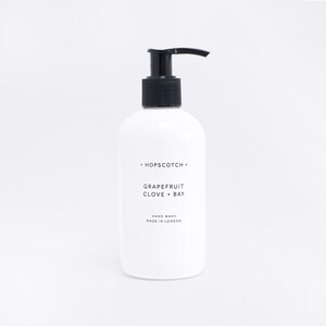 May include: White bottle of hand wash with a black pump dispenser. The label reads "HOPSCOTCH", "GRAPEFRUIT CLOVE + BAY", "HAND WASH", and "MADE IN LONDON".
