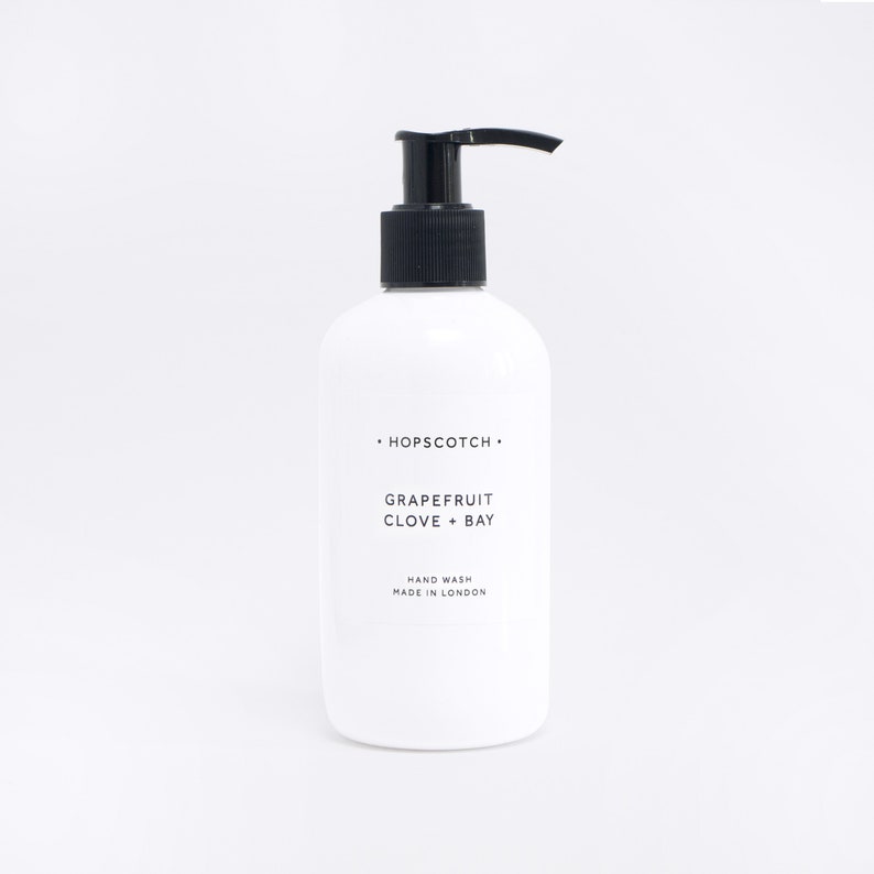 May include: White bottle of hand wash with a black pump dispenser. The label reads "HOPSCOTCH", "GRAPEFRUIT CLOVE + BAY", "HAND WASH", and "MADE IN LONDON".