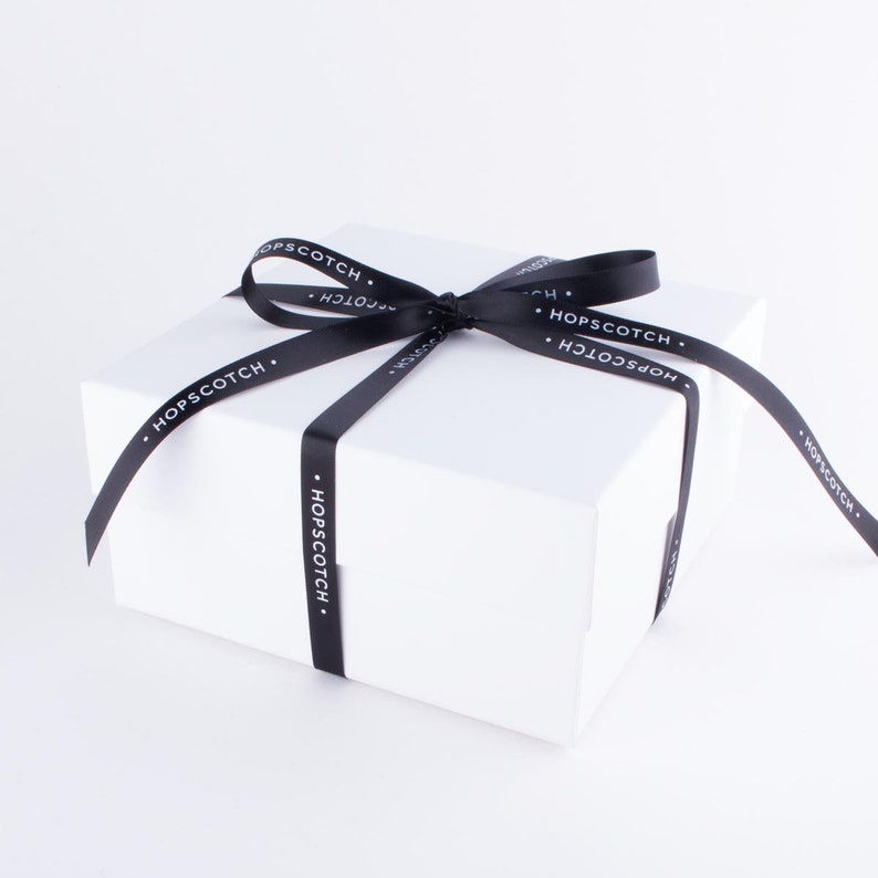 May include: A white gift box with a black ribbon tied in a bow. The ribbon has the text "HOPSCOTCH" printed on it.