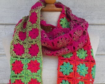 May include: A crocheted scarf with a bright green, orange, and pink floral pattern. The scarf is made of yarn and is displayed on a wooden mannequin.