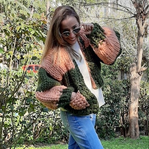 Op de afbeelding: Een vrouw draagt een grove gebreide cardigan met roze en groene strepen. De cardigan is oversized en heeft een ontspannen pasvorm. Ze draagt een blauwe jeans en blauwe sneakers.