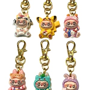 May include: A collection of seven keychains with gold-colored clasps. Each keychain features a unique, small figurine in various colors, including yellow, pink, blue, and white. The figurines have a cartoonish style with exaggerated features and a slightly unsettling expression.